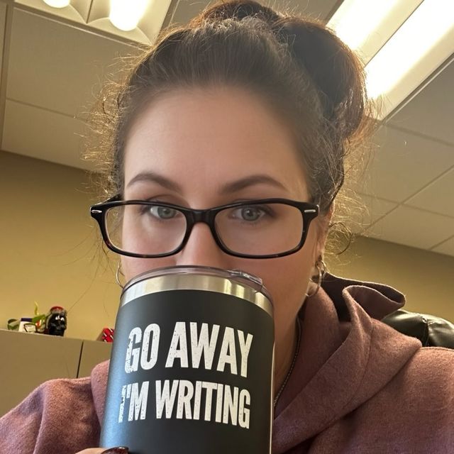 Person with glasses holding a coffee mug that says 'GO AWAY I'M WRITING' in an office setting.